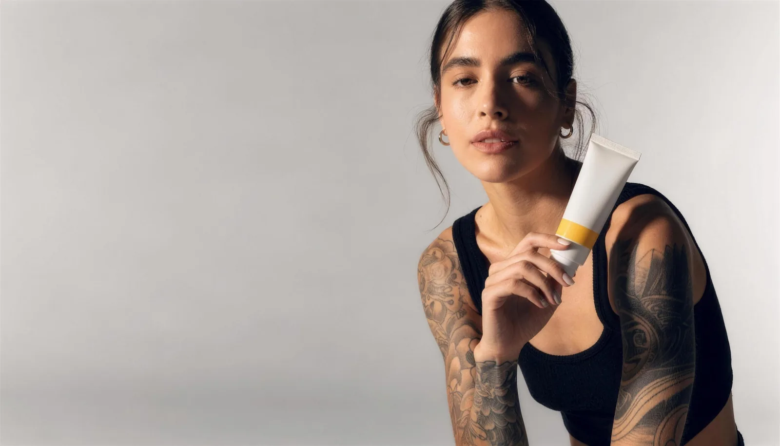 A tattooed woman with black-and-grey sleeve tattoos holding a tube of STNC numbing cream